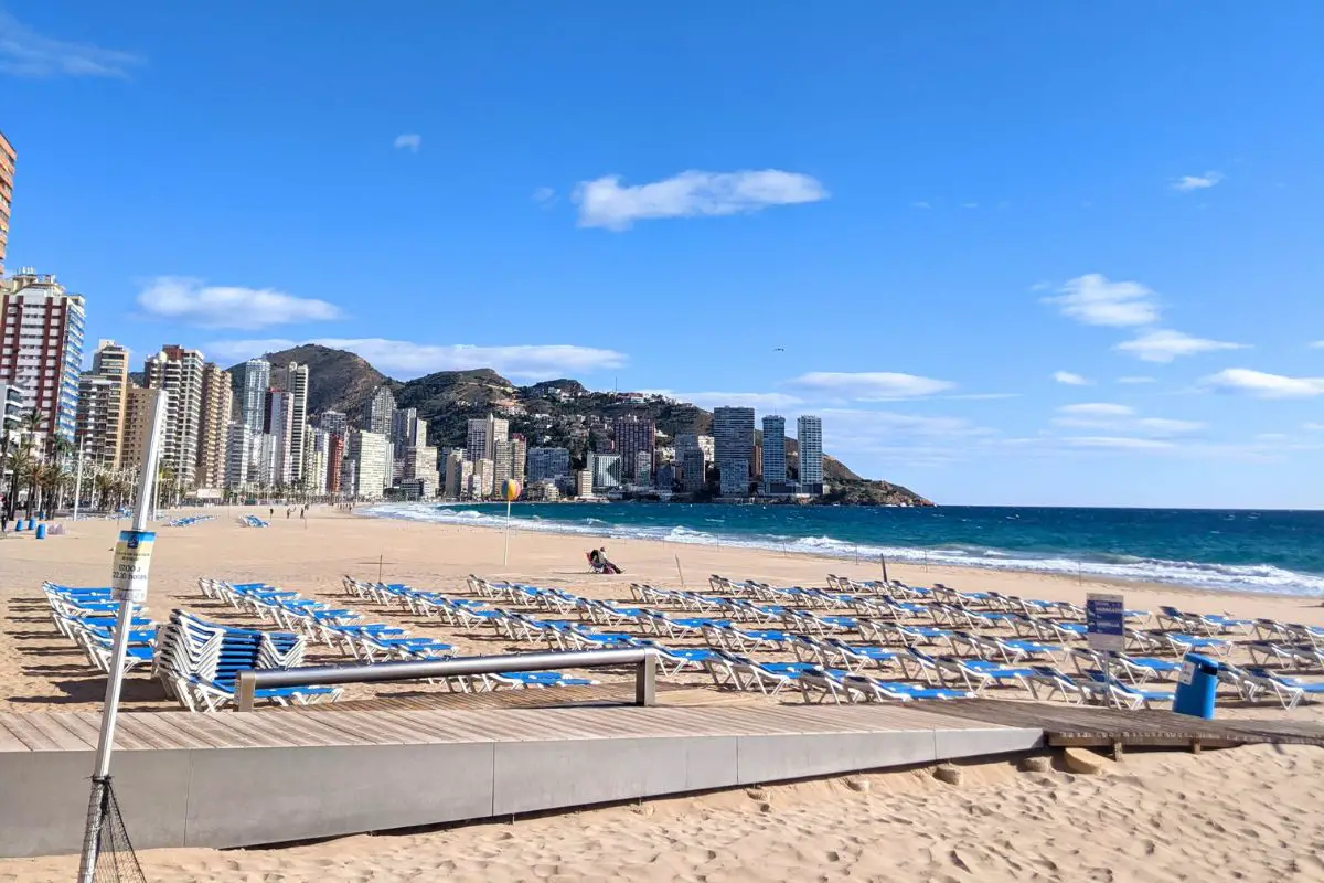 The goldden sandy beach in Benidorm, with a wooden walk way on the sand, blue sunbeds lined up, and high rise buildings and mountains in the background