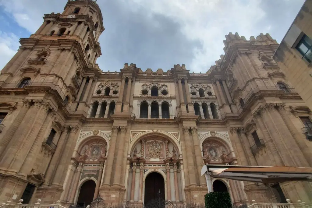 The image shows a view looking up at Malaga Cathedral. The sky is blue with a few clouds.