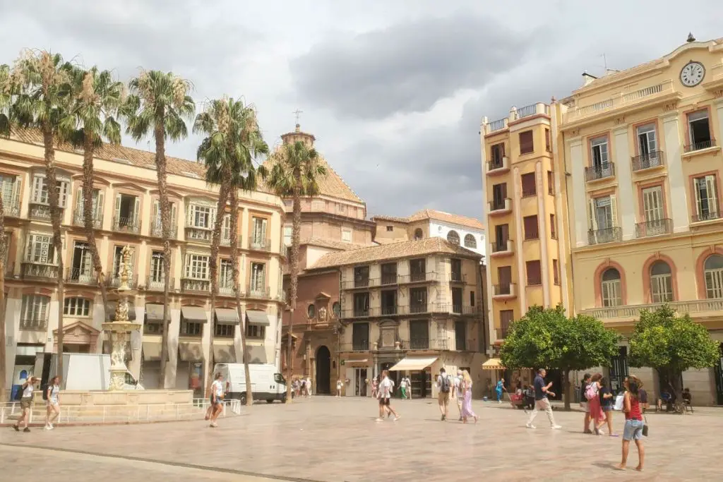 The image shows a plaza in Málaga old town. The tiled square s surrounded by old buildings and palm trees with people walking across the plaza. There are some light grey clouds in the sky.