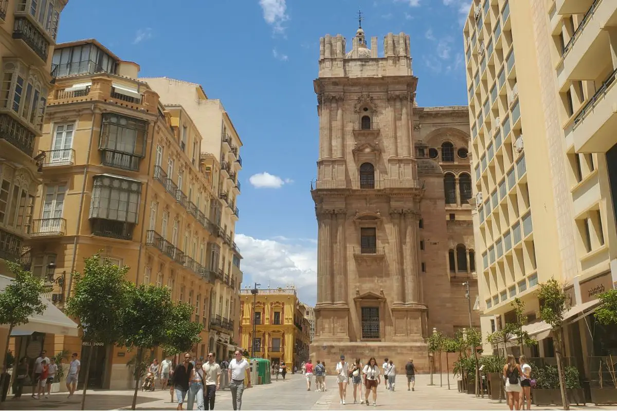 The image shows people walking down a street in Malaga old town. In the centre is the Malaga Cathedral with old buildings to the left and right of it. The sky is blue with a few white clouds.