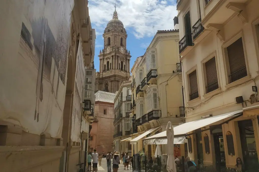 The image shows people walking down a narrow street in Malaga old town. On the right are tables and chairs in front of bars and cafes, and at the end of the street you can see Malaga cathedral.