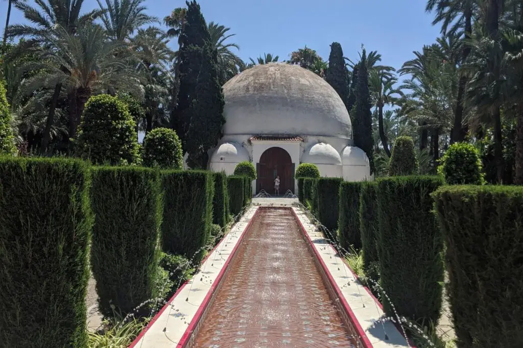 A long pool with fountains, lined with pruned trees, leading up to a white domed building. Located in Elche.