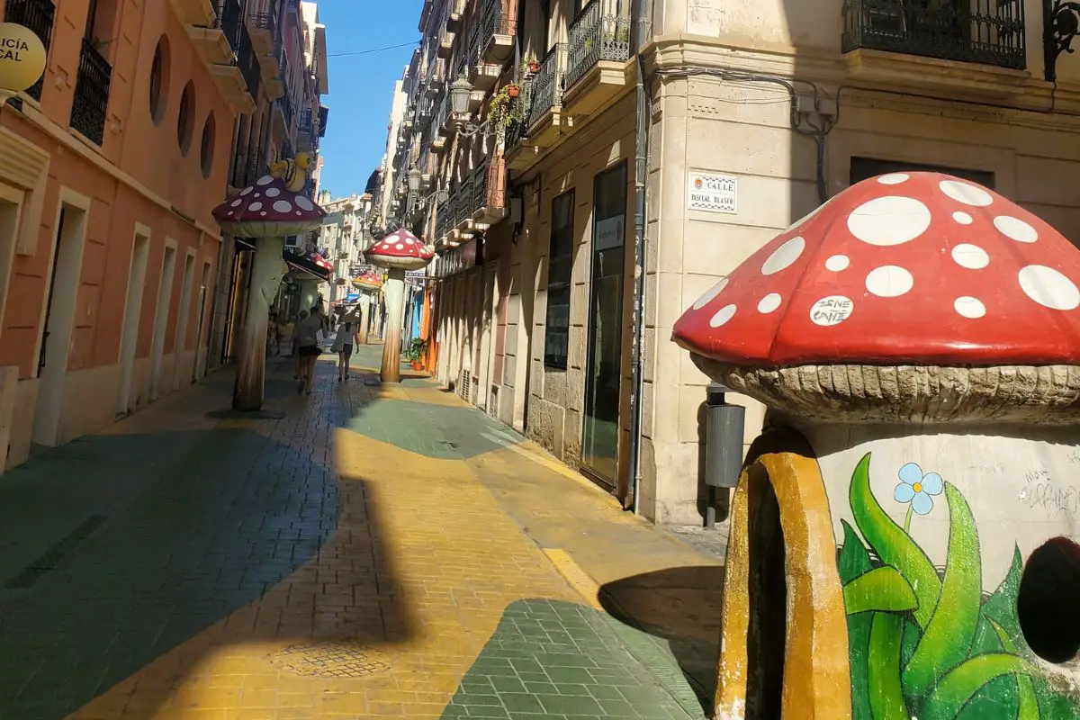 A view down Mushroom Street in alicante. The ground is paved yellow and green, with red and white toadstool playhouses and sculptures along the narrow street.