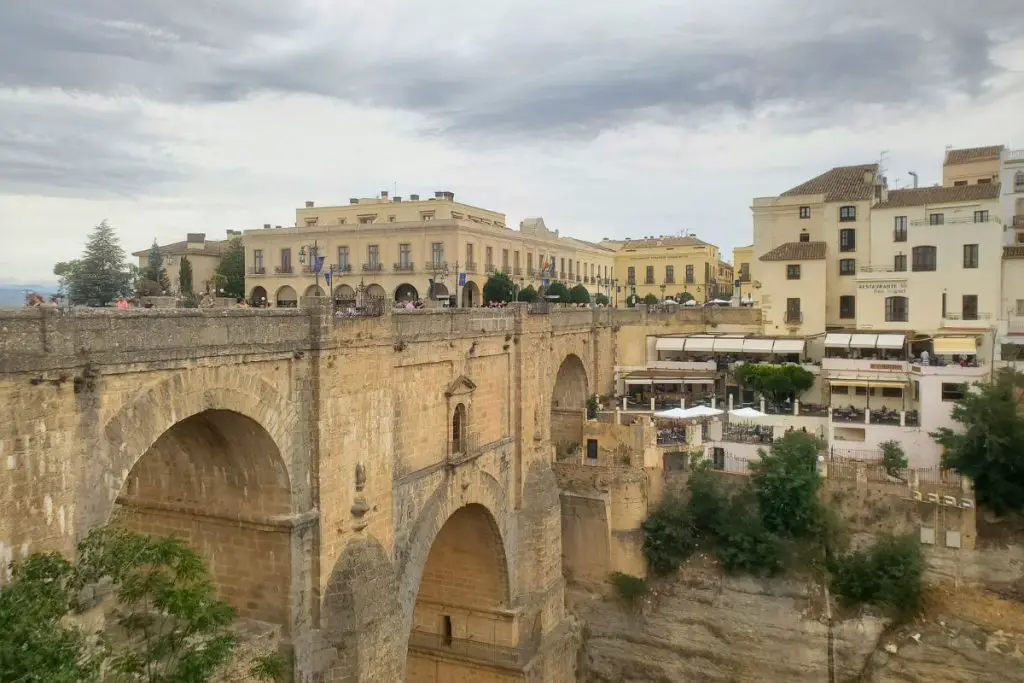 The image shows the view across the El Tajo Gorge towards the Puente Nuevo bridge in Ronda. There are old buildings either side of the bridge and grey clouds in the sky.