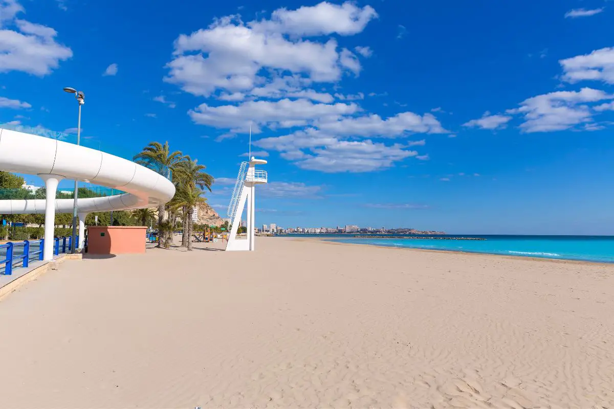 A view of Playa del Postiguet beach in Alicante. Golen sand, turquoise sea, and a white curved bridge and white lifeguard tower.
